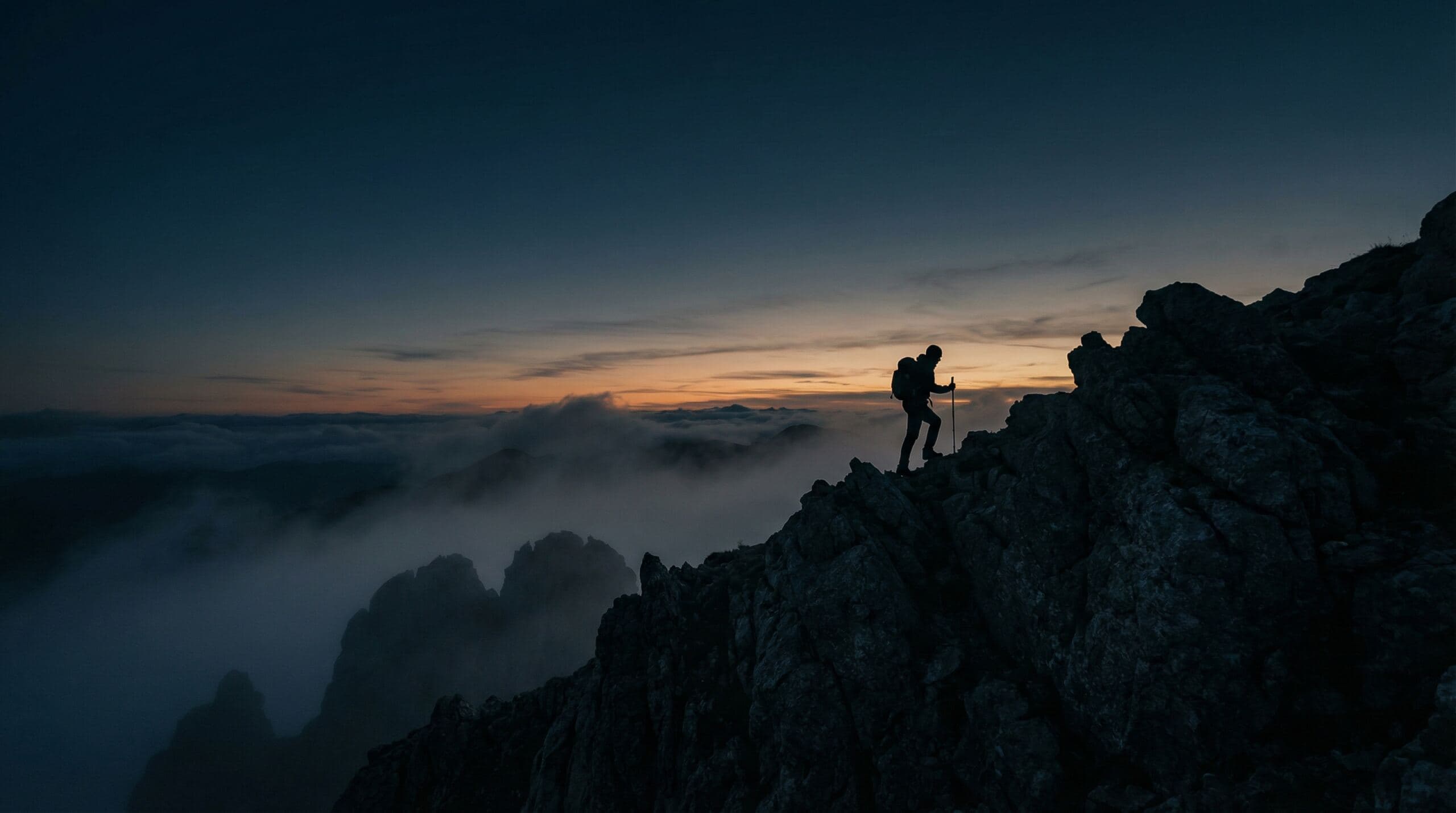 A lone hiker with a trekking pole ascending a rocky ridge above the clouds at dusk